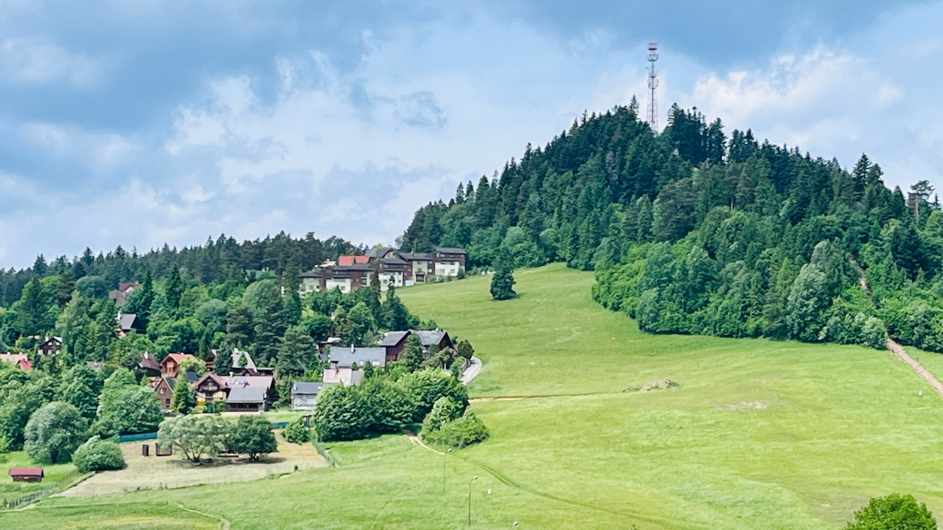 Zelené lúky a lesnatý kopec s domami v údolí, na vrchole stožiar a modrá obloha.